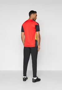 Športový top v červenej, čiernej a žltej farbe, s krátkymi rukávmi a okrúhlym výstrihom. Kombinovaný s čiernymi športovými nohavicami a čiernou obuvou.