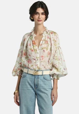 Kobieta ubrana w białą kwiecistą bluzkę z falbanianym kołnierzem i bufkami na rękawach, jasnoniebieskie dżinsy, kremowy pasek ze złotą klamrą oraz złote kolczyki.