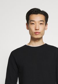 T-shirt long noir à manches longues avec un col rond et un col intérieur blanc, fabriqué en tissu côtelé, présentant une coupe ajustée et un design minimaliste.
