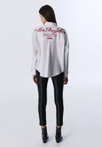 Camisa blanca a rayas con texto rojo "Los Ángeles" en la parte de atrás, con dobladillo redondeado, combinada con leggings de cuero sintético negros y botas de tobillo.