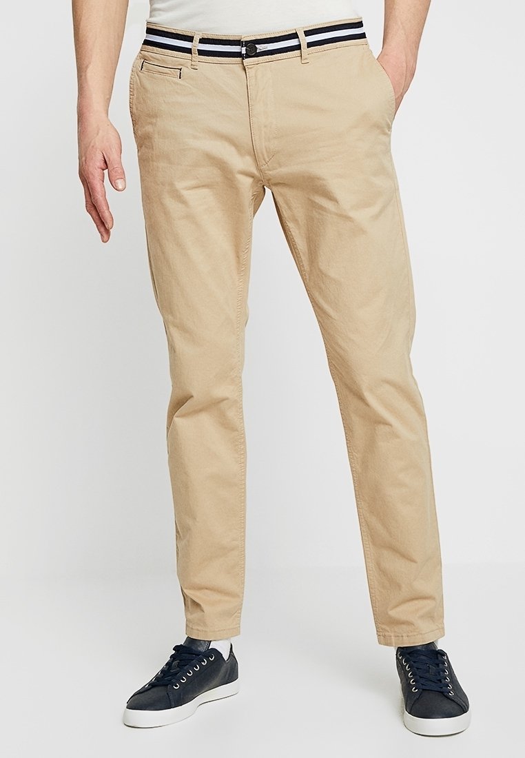 Béžové chino kalhoty se slim fit střihem, s černobílým pruhovaným pasem, předními kapsami a hladkou bavlněnou texturou. Oblečené s námořnickými teniskami.