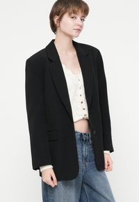 Černý oversized blazer s klopovým límcem, zapínáním na jeden knoflík a přední kapsou. Nosit s krémovým cropped topem a světle modrými džíny.