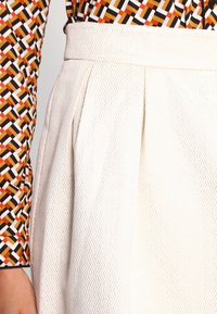 Valkoinen teksturoitu hame, jossa on poimutuksia, yhdistettynä kuvioituun pitkähihaiseen puseroon, jossa on mustia, valkoisia, oransseja ja keltaisia geometrisia muotoja.