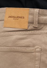 Gros plan sur un pantalon beige montrant une poche arrière et une étiquette en cuir tanné cousue avec le texte « JACK & JONES DEPUIS 1990 ».