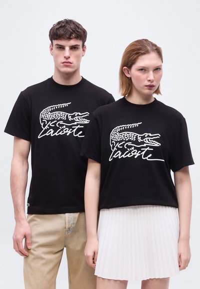 Млад мъж и жена облечени с черни тениски на Lacoste с бяло лого крокодил; мъжът е с бежови панталони, жената с бяла плисирана пола, неутрален фон.