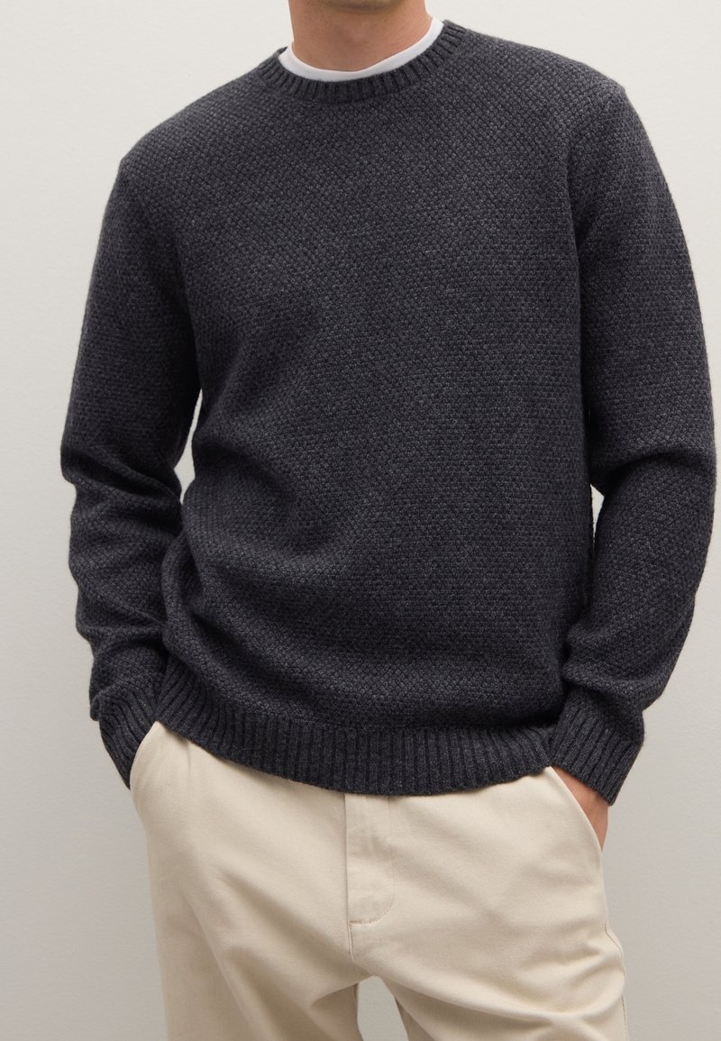 Homme portant un pull en maille texturée gris foncé sur une chemise blanche, les mains dans les poches d'un pantalon beige, devant un fond uni.