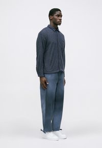 Μπλε πουκάμισο υφασμάτινο με κουμπιά και μακριά μανίκια, σε συνδυασμό με ανοιχτό μπλε παντελόνι φαρδιάς γραμμής και λευκά αθλητικά παπούτσια. Απλός σχεδιασμός, χωρίς ορατά σχέδια.