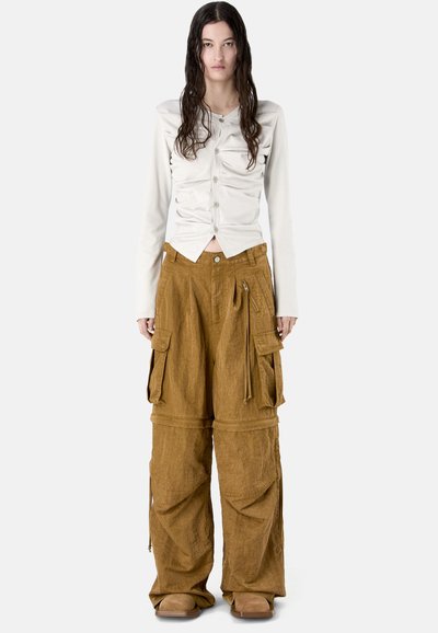 Kobieta o długich ciemnych włosach nosi białą marszczoną bluzkę z guzikami oraz oversize'owe brązowe spodnie typu cargo z dużymi kieszeniami i beżowe buty.