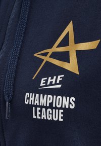 Navyblauwe rits-hoodie met een gestructureerd gouden logo en witte tekst, gemarkeerd met "EHF CHAMPIONS LEAGUE," en een trekkoord op de kap.