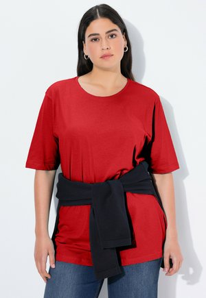 T-shirt in cotone rosso con maniche corte e scollatura rotonda; maglione blu navy legato in vita; il modello indossa jeans blu. Design semplice e informale.