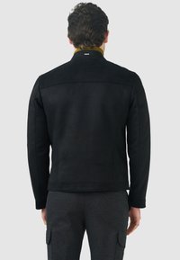 Giacca in suede nera con design strutturato, caratterizzata da un colletto a coste giallo e dettagli minimalisti sulla schiena, vestibilità slim e texture liscia.