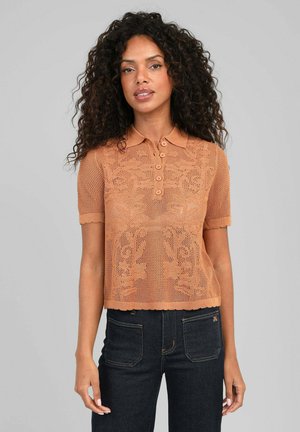 Femme aux cheveux bouclés portant un polo en maille marron à manches courtes avec des motifs floraux et un jean sombre taille haute.