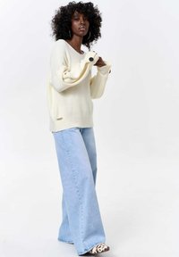 Oversized cream sweater with ribbed texture and black button accents, paired with light blue wide-leg jeans and patterned shoes.