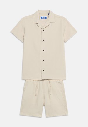 Beige kortærmet skjorte med knapper og reverskrave kombineret med matchende shorts med snøre, lagt fladt på en hvid baggrund.