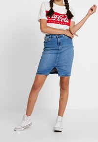 Lichtblauwe denim kokerrok met een voorkruis, hoge taille en t-shirt met logo. Gecoupeerd met witte hoge sneakers.