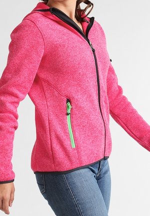 Mujer con chaqueta rosa brillante con cremallera verde y pantalones vaqueros azules, de pie con los brazos ligeramente extendidos.