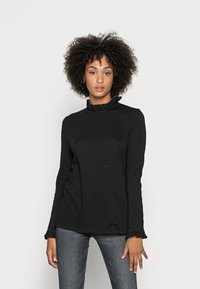 Zwart langemouwen top met een ronde, gerimpelde halslijn en subtiele textuur. Strakke mouwen met ruches aan de manchetten. Jeans zichtbaar daaronder.