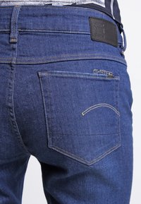 Modré džíny z denimu s kapsou na zadní straně se šitým vzorem, viditelnou značkovou štítkem a hladkou texturou. Pas má poutka na pásek.