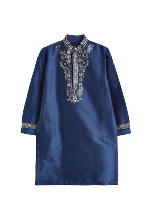 Niño pequeño sonriendo, vestido con una túnica tradicional azul bordada con pantalones blancos y sandalias azules, de pie frente a un fondo beige.