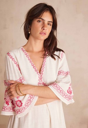 Jonge vrouw met bruin haar, gekleed in een witte jurk met roze borduursels en gouden armbanden, staand met gekruiste armen tegen een effen achtergrond.