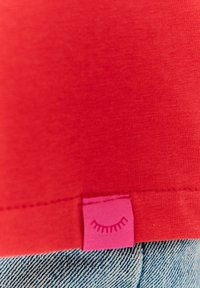 Czerwona bawełniana koszulka z małą różową metką z prostym wzorem oka, umieszczona w pobliżu dołu, wyeksponowana na niebieskim dżinsowym materiale.