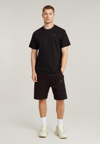 Completo in cotone nero composto da t-shirt e pantaloni corti, con un piccolo logo sul petto. Modello che indossa sneaker chiare e calzini bianchi alla caviglia.