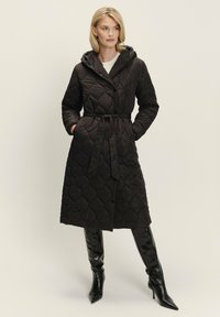 Čierny prešívaný dlhý kabát s kapucňou, opaskom v páse a diamantom vzorom. Kombinovanie s lesklými čiernymi vysokými čižmami.