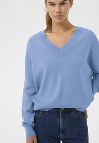 Világoskék V-nyakú pulóver puha anyagból, eresztett vállakkal és bordázott mandzsettákkal, magas derekú sötétkék farmernadrággal párosítva.