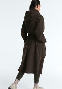 Kvinde iført en mørkebrun trenchcoat med hætte, sorte leggings, lyserøde sokker og hvide sneakers, stående med hænderne i frakkens lommer.