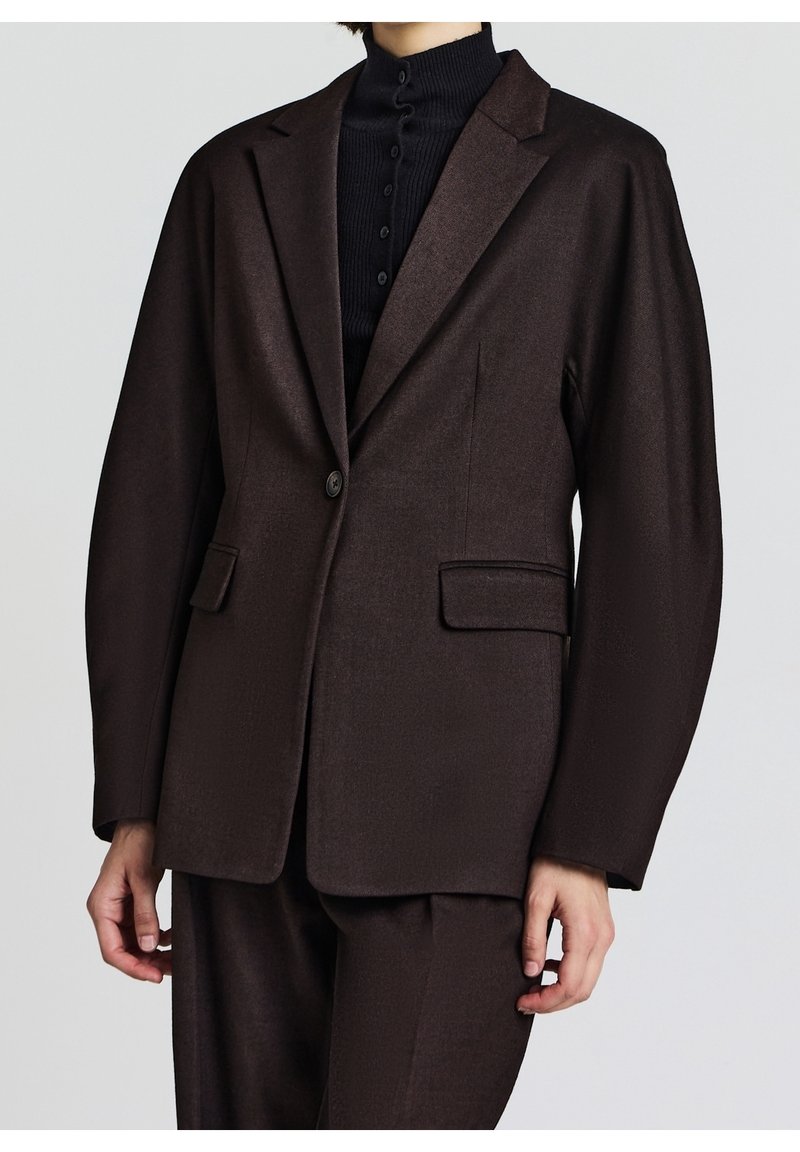 Personne portant un blazer marron foncé ajusté avec des poches à rabat sur un pull à col roulé côtelé noir boutonné.