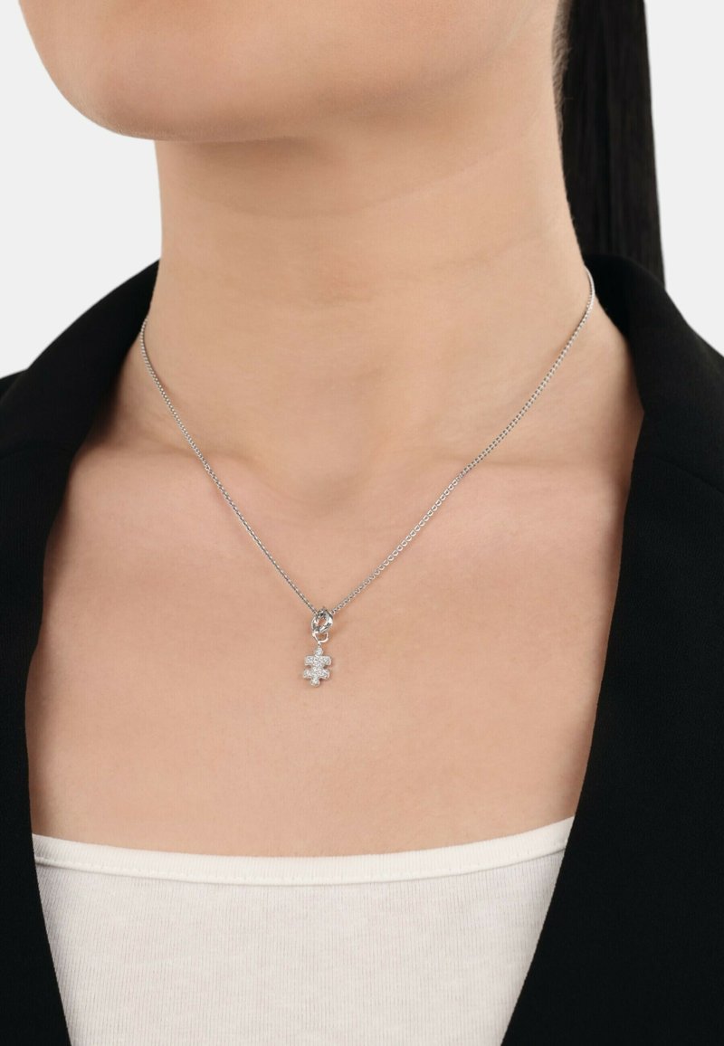 Silberkette mit einem kleinen Kleeblatt-Anhänger, verziert mit funkelnden Akzenten, getragen auf nackter Haut über einem hellen Oberteil.