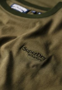 Oliivivihreä puuvillapaita, jossa on ribbikaarroke, ja tummemman oliivinvärinen kirjailtu teksti "SuperDry Athletics". Kirjoitettava pinta.