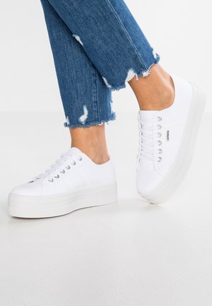 White canvas sneakers with a thick sole, round toe, and five metallic eyelets for laces. Worn with distressed blue denim jeans.