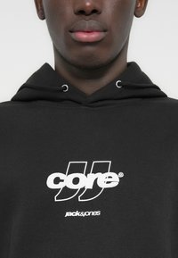 Κοντινό πλάνο ενός ατόμου που φοράει μαύρο φούτερ με κουκούλα με λευκά λογότυπα κειμένου "core" και "jack & jones" στο στήθος.