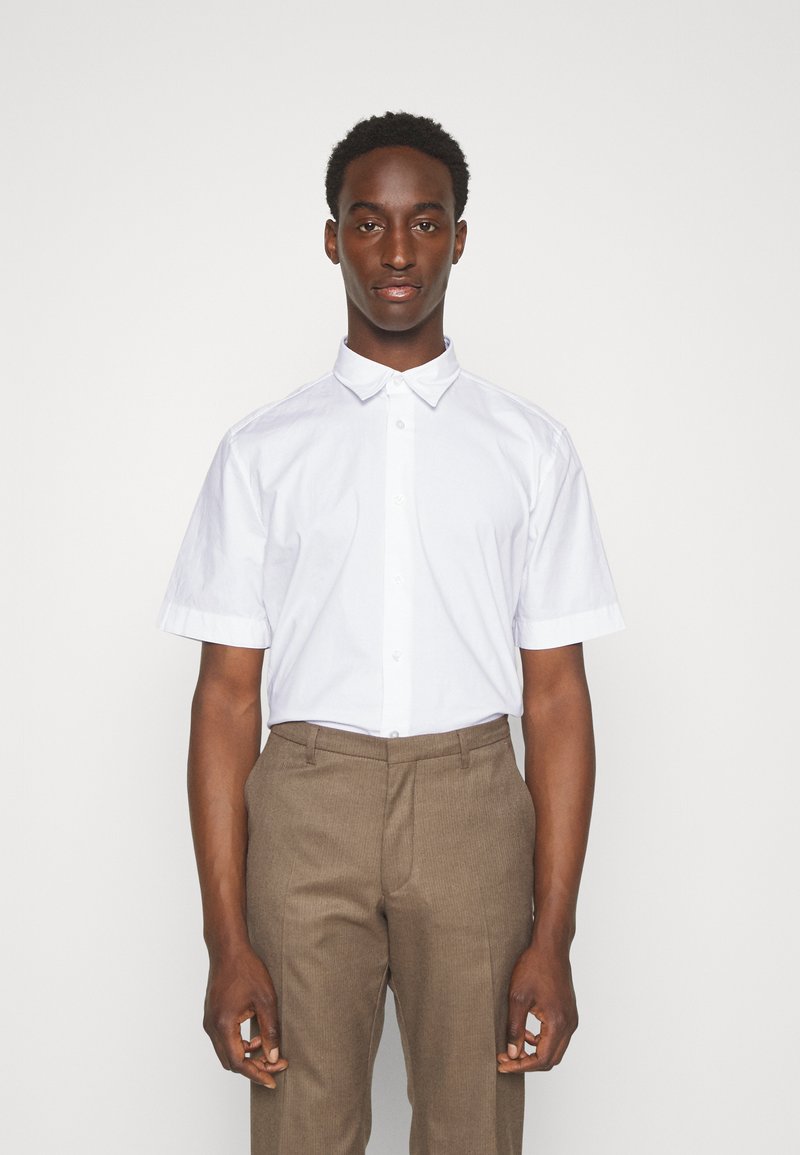 Camisa blanca de manga corta con cuello estructurado, textura lisa y botones al frente. Combinada con pantalones marrones a medida, que presentan un patrón sutil.
