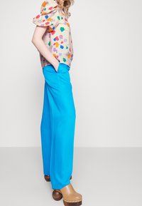 Bloemige blouse met puffmouwen in pasteltinten, gecombineerd met felblauwe wijde broek en houten klompen met metallic accenten.