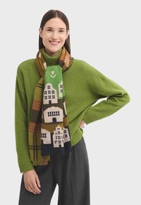 Pull en tricot vert, écharpe à motifs avec des maisons et des arbres, couleurs noir, marron, vert et jaune, associée à un pantalon noir à rayures.