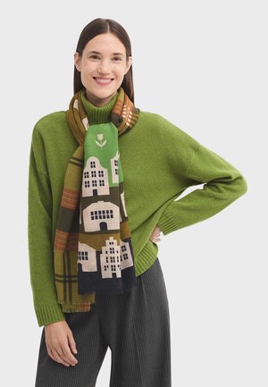 Maglione verde a maglia, sciarpa con motivo di case e alberi, colori nero, marrone, verde e giallo, abbinato a pantaloni scuri a righe verticali.