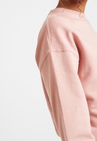 Růžová bavlněná mikina s kulatým výstřihem, žebrovanými manžetami a volným střihem. Výrazné švy jsou viditelné podél ramen a rukávů.