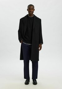 Ilgas juodas vilnonis paltas ant tamsaus megztinio, derinamas su pritaikytais mėlynais kelnais ir juodais batais. Yra klasikinis apykaklė ir šoninės kišenės.