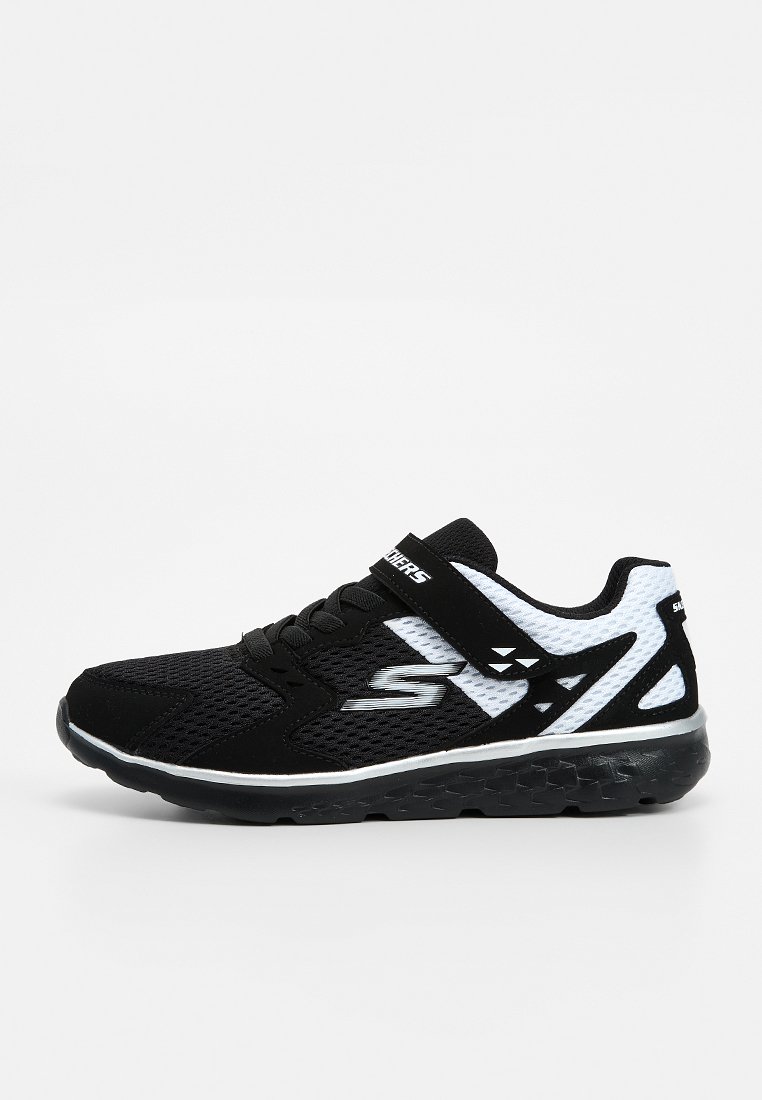 Zapatilla deportiva Skechers en blanco y negro con parte superior de malla, cordones en la parte delantera y tira de Velcro sobre un fondo liso.