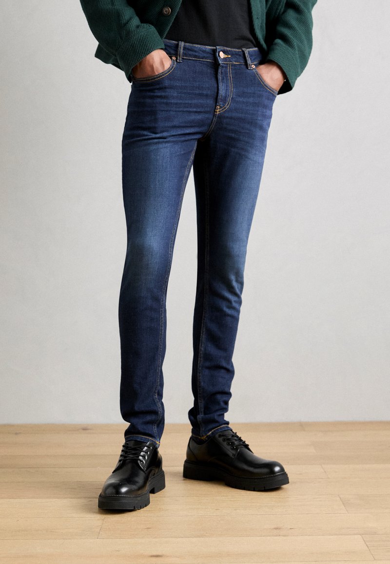 Pantalones vaqueros de denim azul oscuro con un corte ajustado, que cuentan con costuras doradas, un diseño clásico de cinco bolsillos y un patrón de costura visible a lo largo de las costuras.
