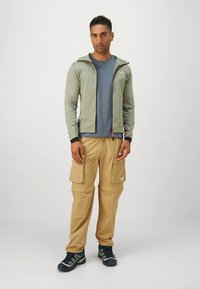 Chaqueta deportiva gris con cremallera sobre una camiseta azul, combinada con pantalones cargo color beige con bolsillos. Zapatillas azules completan el conjunto.