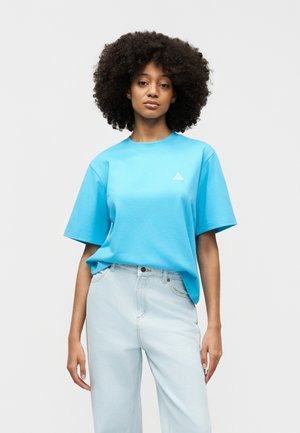 Ung kvinde med krøllet hår iført en stor, knaldblå t-shirt og lyseblå højtaljede jeans, stående foran en ensfarvet baggrund.