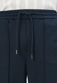 Dunkelblaue Hose mit einem elastischen Bund und verstellbarem Kordelzug mit silbernen Akzenten. Glatte Textur und taillierte Passform.