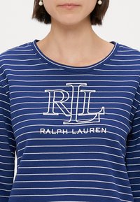 Μπλε ριγέ μπλούζα με μακριά μανίκια, διακοσμημένη με λευκές οριζόντιες ρίγες, στρογγυλή λαιμόκοψη και κεντημένο λογότυπο "RL" με το κείμενο "RALPH LAUREN" από κάτω.