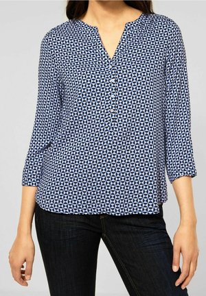 Mujer con una blusa de patrón geométrico azul y blanco con mangas tres cuartos y pantalones oscuros, mostrada desde el cuello hasta las caderas.