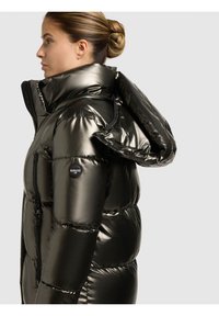 Pūdera jaka tumši metāliskā zaļā krāsā ar augstu apkakli, rāvējslēdži priekšpusē un logotipa uzlikas uz piedurknes. Dizainam ir spīdīga tekstūra.