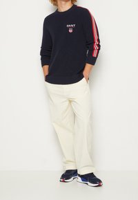 Person in einem dunkelblauen Pullover mit roten und weißen Streifen an den Ärmeln, cremefarbenen Hosen und dunkelblauen Sportschuhen mit roten Akzenten.