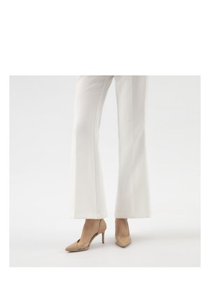 Persoon draagt witte broek met wijde pijpen en beige puntige hoge hakken, staand op een effen witte achtergrond.
